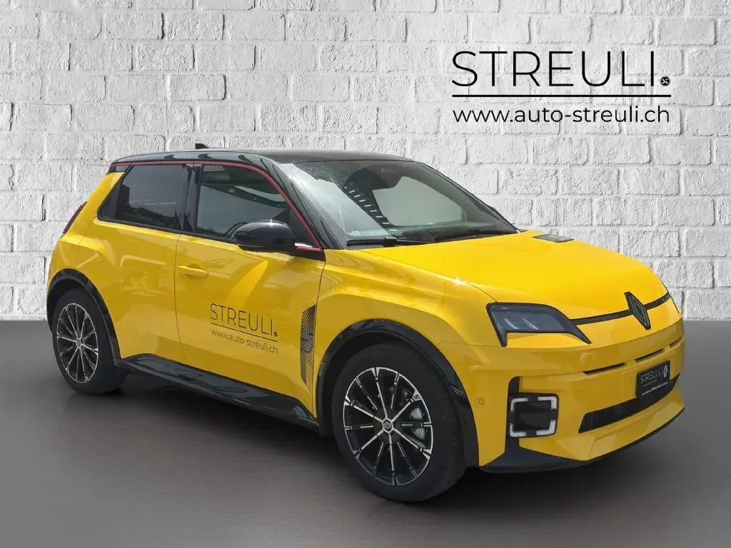 Schrägansicht von vorne rechts eines gelben Renault 5 E-Tech Electric mit schwarzem Dach vor einer weissen Ziegelwand.